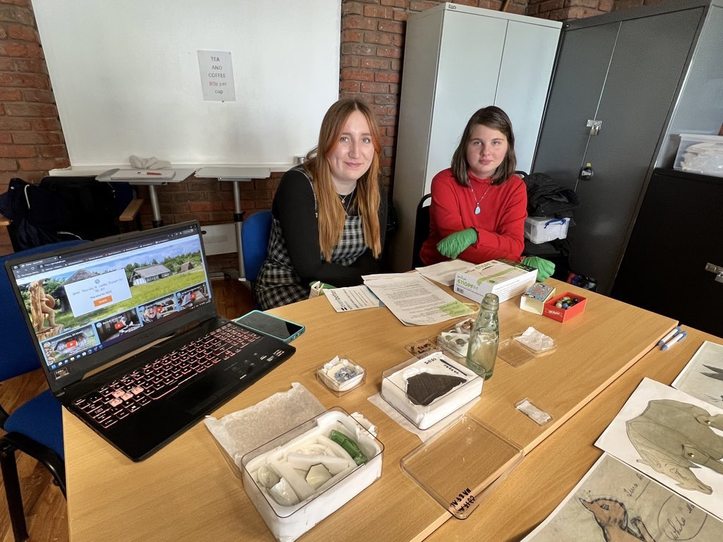 Two of our volunteers (Amy and Maria) sat at a table covered in items from the collection such as a glass bottle and pieces of broken pottery padded in a storage box. They also have a laptop open showing off the website.